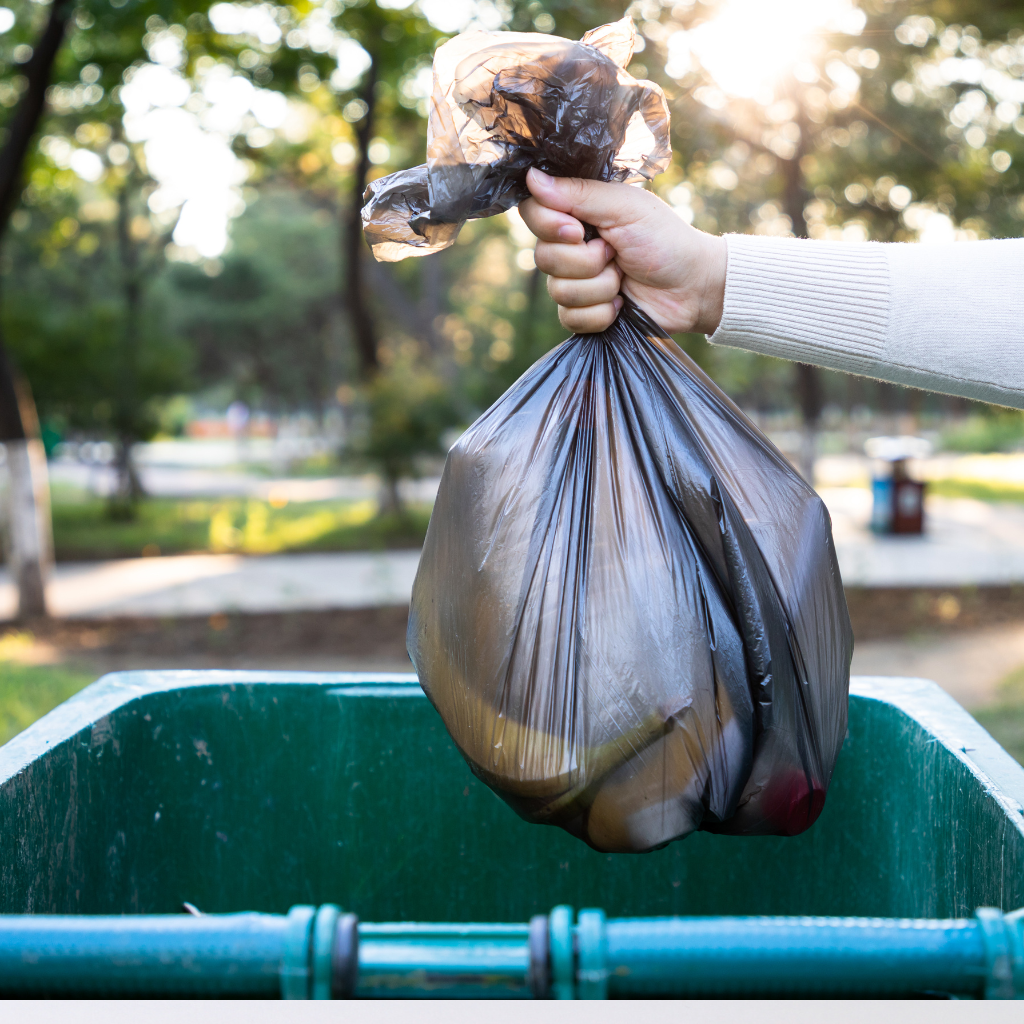 an open green trash can with a arm stretched over it holding a black bag of trash